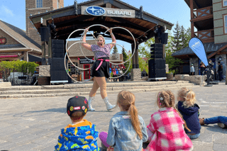 A group of kids sitting on the ground in front of the Subaru Stage as a professional hula hoop performer shows off her skills with 5 hoops on a sunny afternoon.